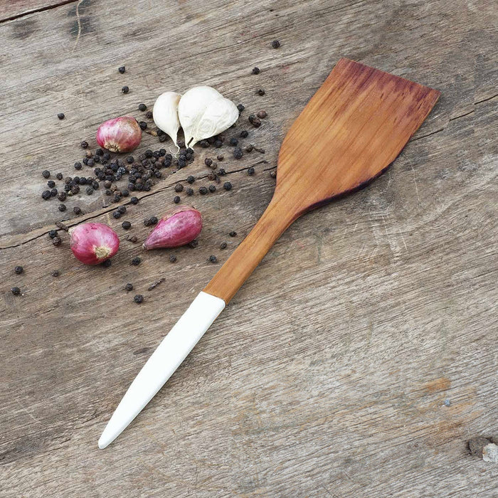 Extra large teak spatula with white handle next to shallots, garlic, and peppercorn displayed on a wooden surface