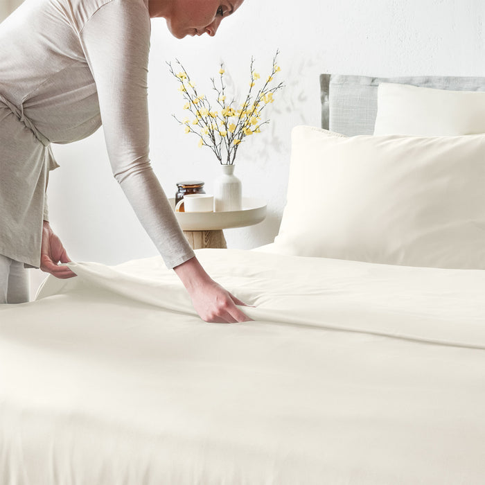 Person making a bed in a bright bedroom with white bedding and decorative elements.
