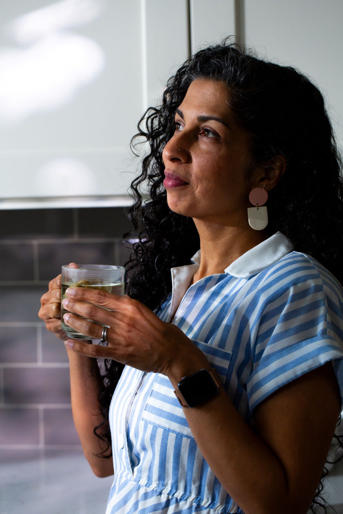 Woman in blue striped shirt holding glass of water, looking thoughtfully toward window in bright indoor setting.
