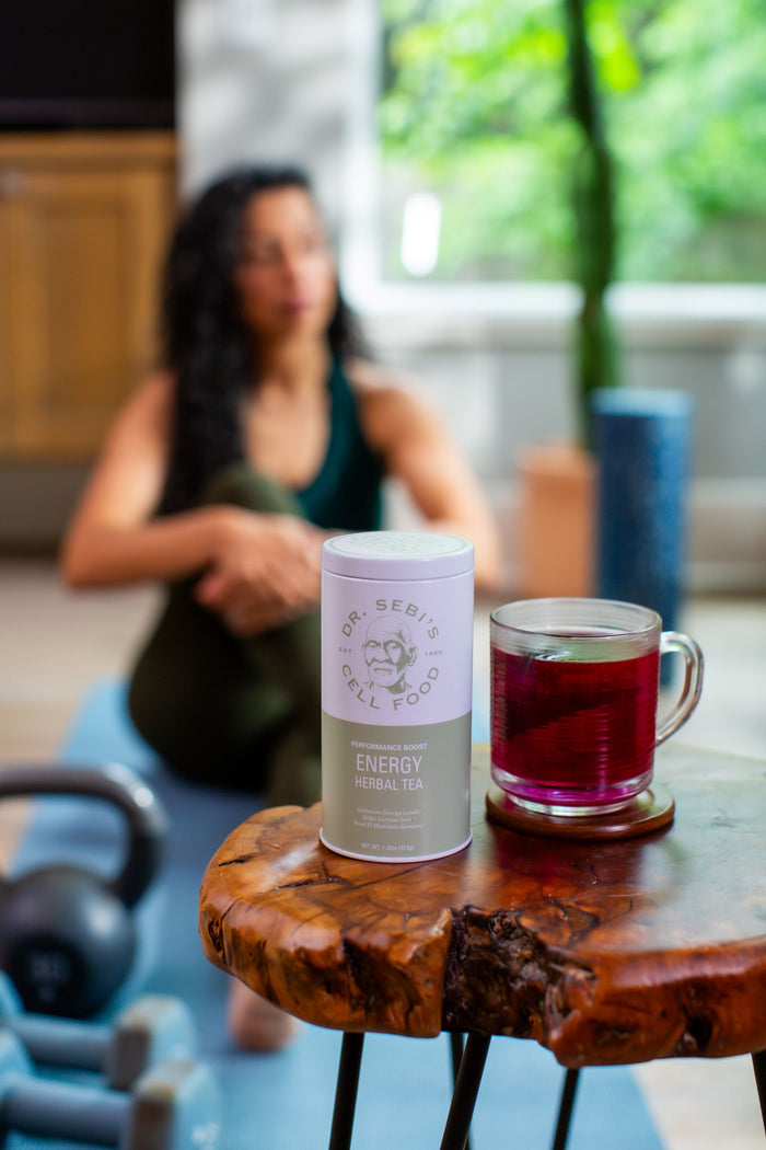 Dr. Sebi's Energy Herbal Tea displayed with brewed cup beside dumbbells in a wellness setting with woman in background.