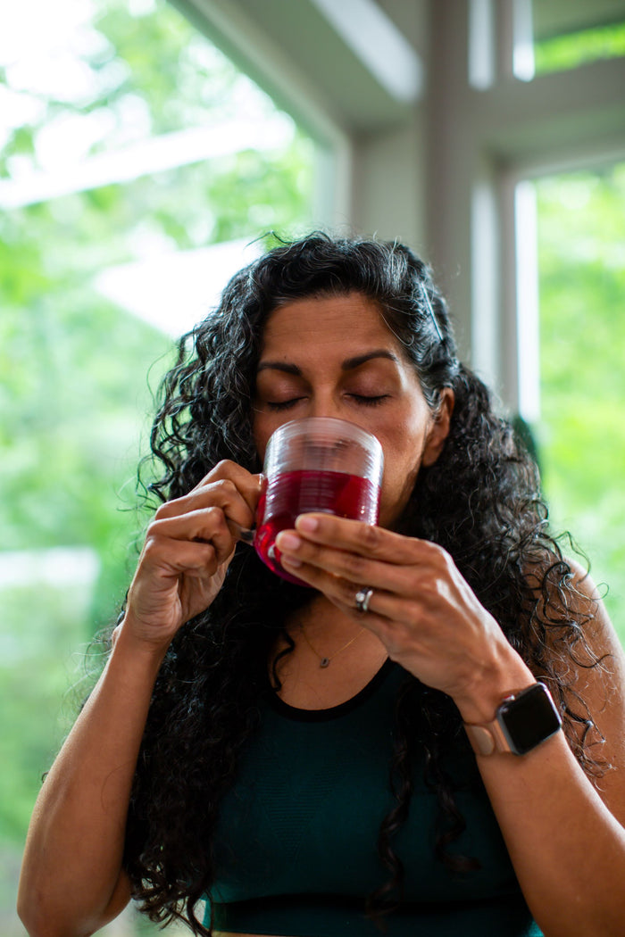 Woman with long curly hair drinking red herbal beverage by a bright window with green foliage in background.