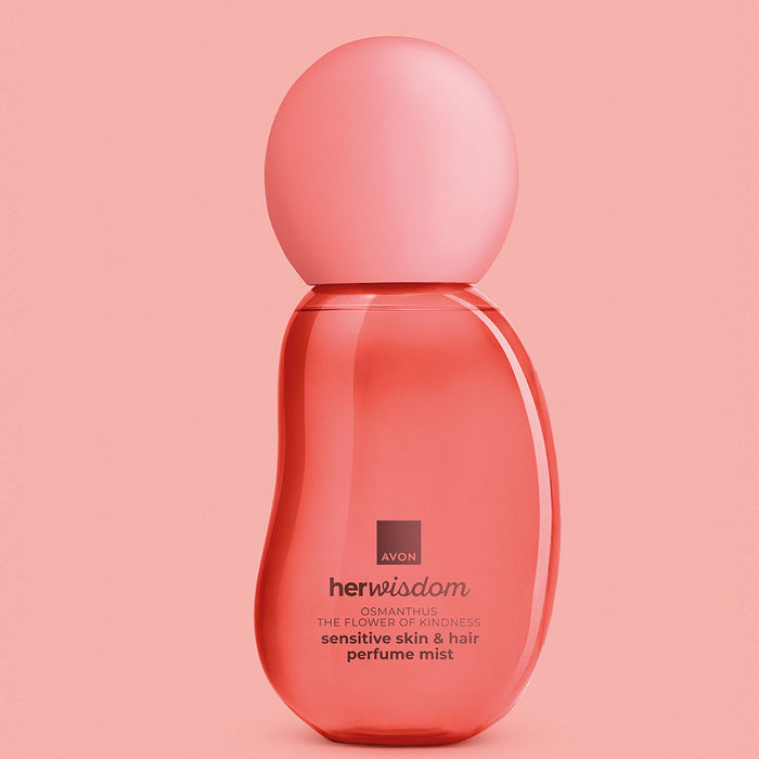 A bottle of Avon Her Wisdom Osmanthus sensitive skin & hair perfume mist in a translucent coral pink color, set against a solid pink background.