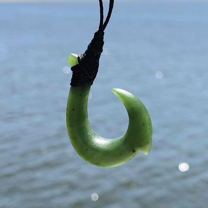 Light green jade hook pendant hanging from a black cord against a blue water background