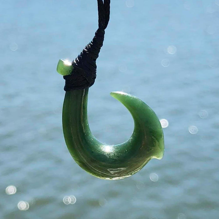 Green jade hook pendant curved into a crescent shape hanging from a black cord against a blue water background