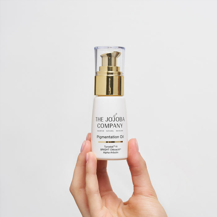 Hand holding a bottle of 'The Jojoba Company' Pigmentation Oil against a light gray background