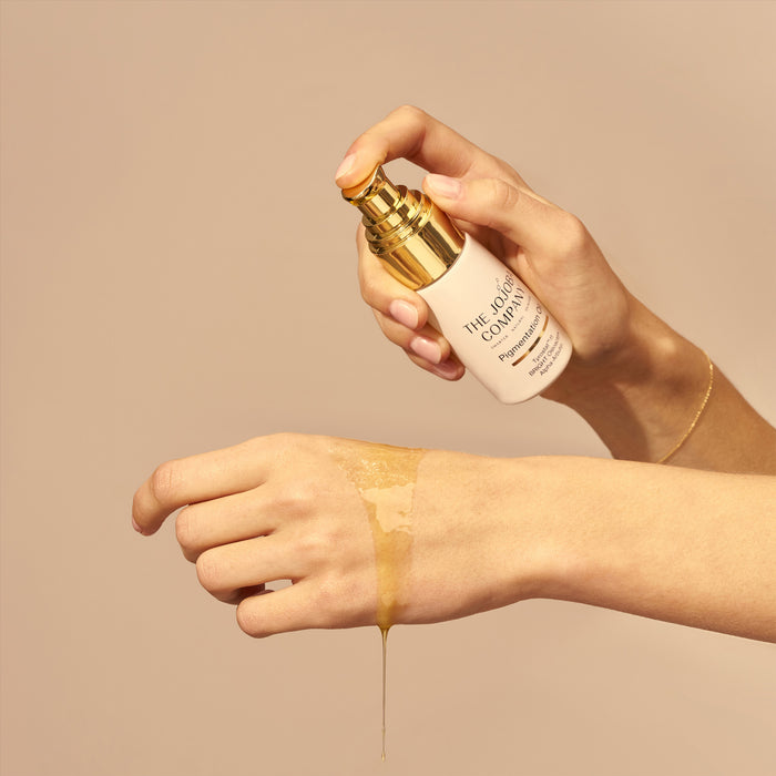 Hand applying a Pigmentation Oil product from 'The Face Company' to a wrist against a beige background