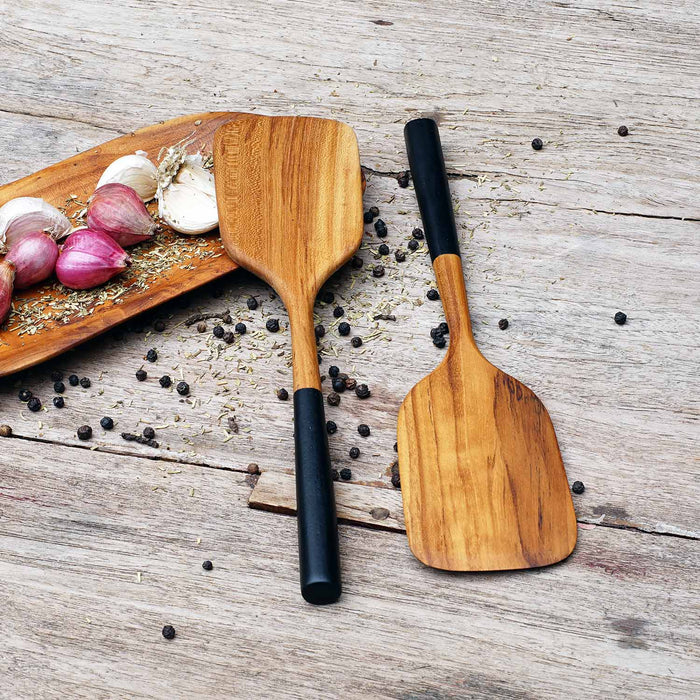 2 Rico & Plato bistro teak spatula with black handles on a wooden plate with shallots, garlic, herbs, and peppercorns on a wooden surface