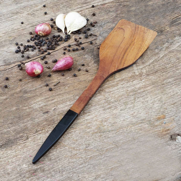 Rico & Plato resto teak spatula with black handle next to peppercorns, shallots, and garlic on a wooden surface