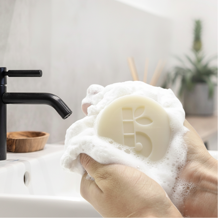 Hand holding a bambaw shaving soap bar of soap with lather, in front of a sink with a black faucet.