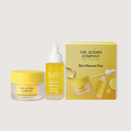 Skincare products from The Jojoba Company including a jar, bottle, and box on a light gray background.