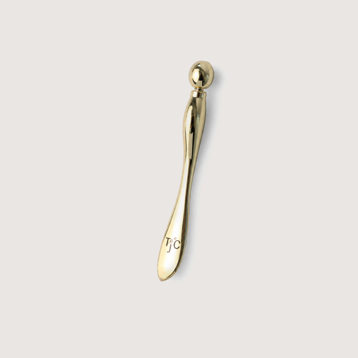 A polished gold facial roller tool with a spherical bead on one end and a smooth curved handle engraved with the initials 'TJC', shown against a solid light gray background.