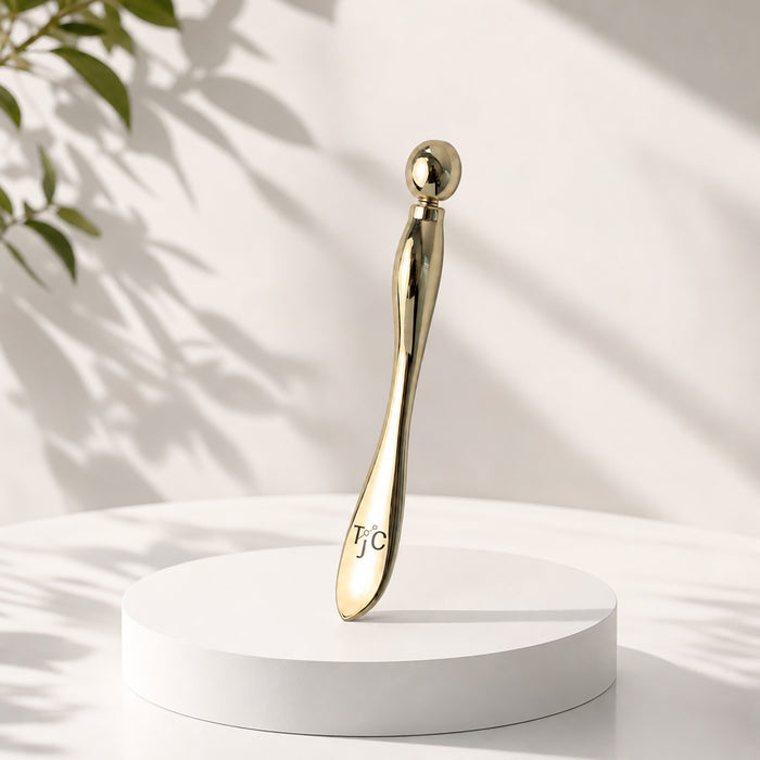 A polished gold facial massage tool with a spherical handle end and a curved body engraved with 'TJC', displayed on a round white pedestal against a softly lit white background with leaf shadows.