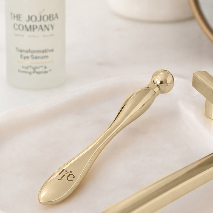 A polished gold facial roller tool with a spherical end and a monogram engraving, resting on a white marble tray next to a bottle of The Jojoba Company Transformative Eye Serum.