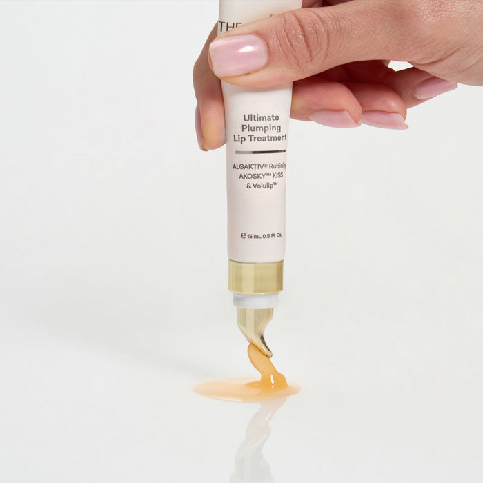 A hand with pale pink manicured nails squeezes a tube of lip treatment, releasing a glossy amber-colored gel onto a clean white surface. The tube is cream-colored with black text reading 'Ultimate Plumping Lip Treatment' and a gold cap.