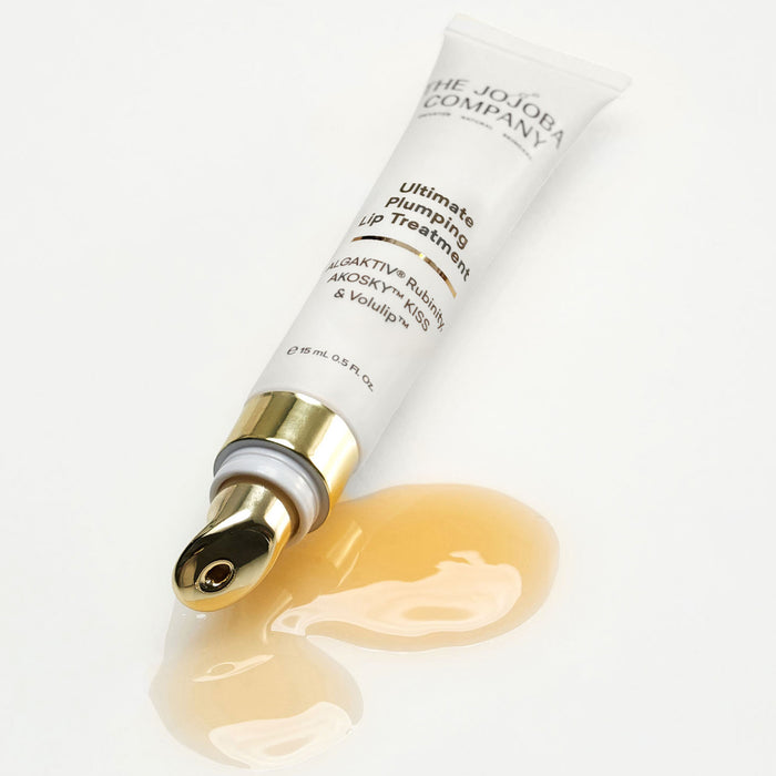 A white squeeze tube of The Jojoba Company Ultimate Plumping Lip Treatment with a gold cap, shown on a white background with a glossy smear of the pale golden treatment below it.