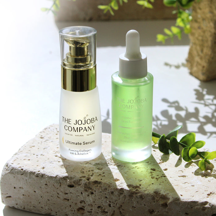 Two skincare products from The Jojoba Company on a stone surface with greenery in the background